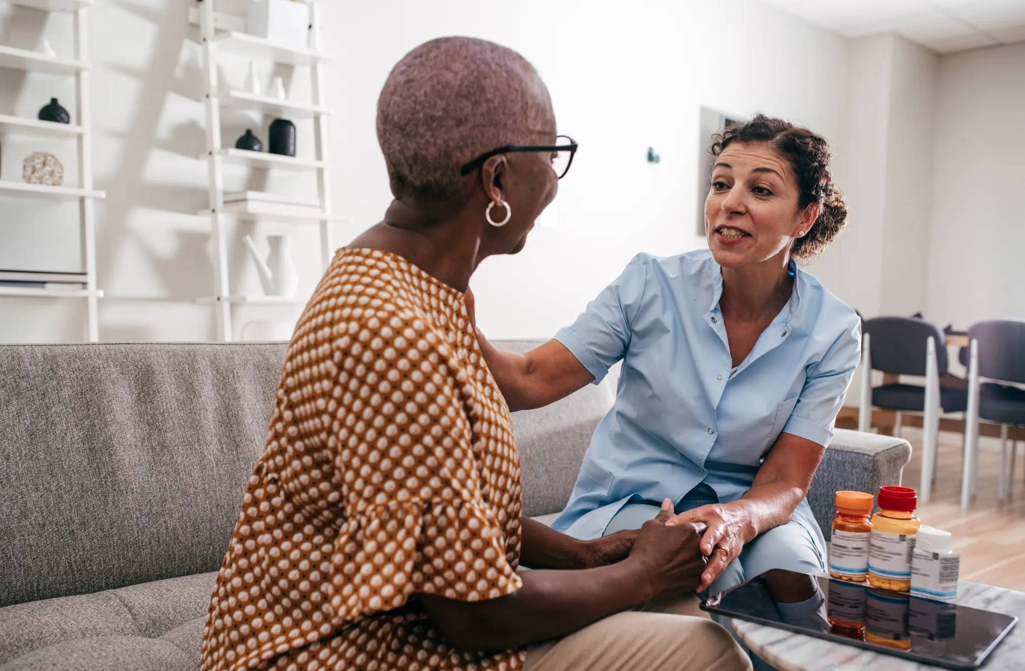 Caregiver in scrubs comforting a senior woman on a living room couch, with medication bottles on the table nearby