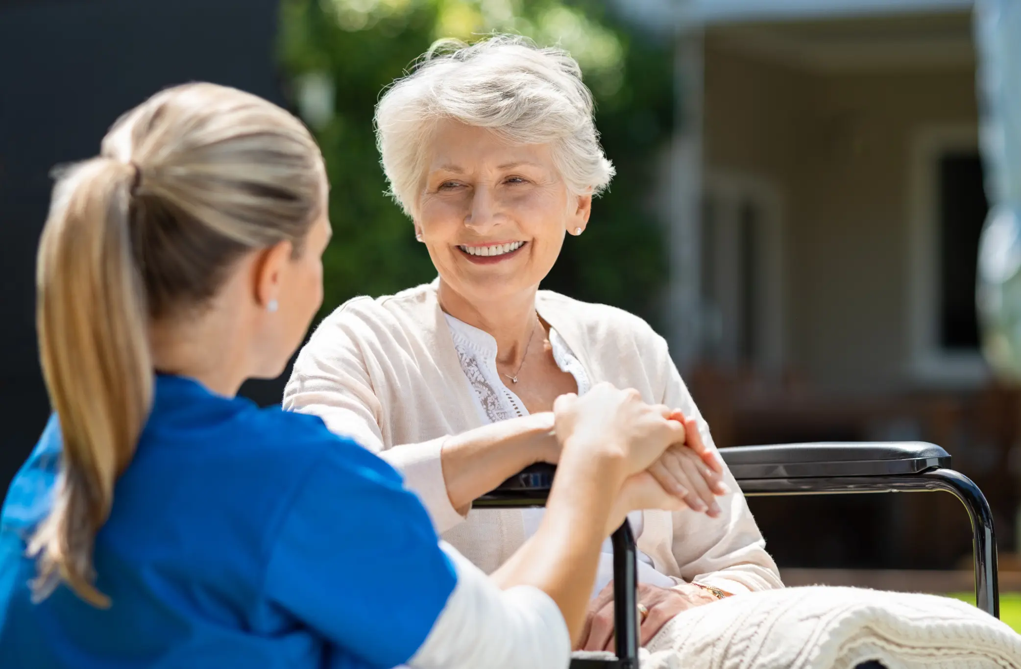 Respite caregiver holding hands with a smiling senior woman in a wheelchair outdoors during an in-home respite care visit